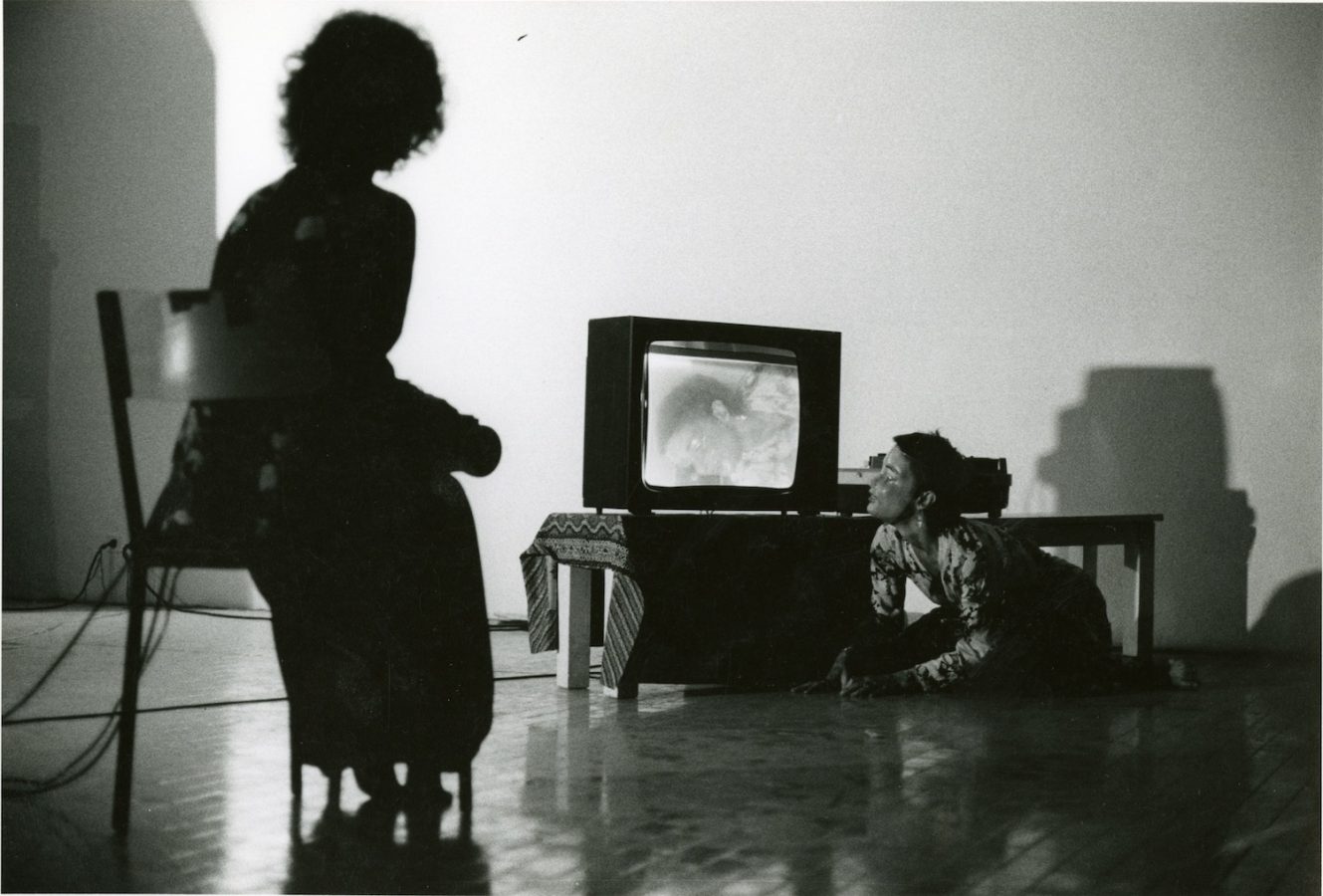 A performer sits in a chair while another crouches on the floor, looking at a video monitor