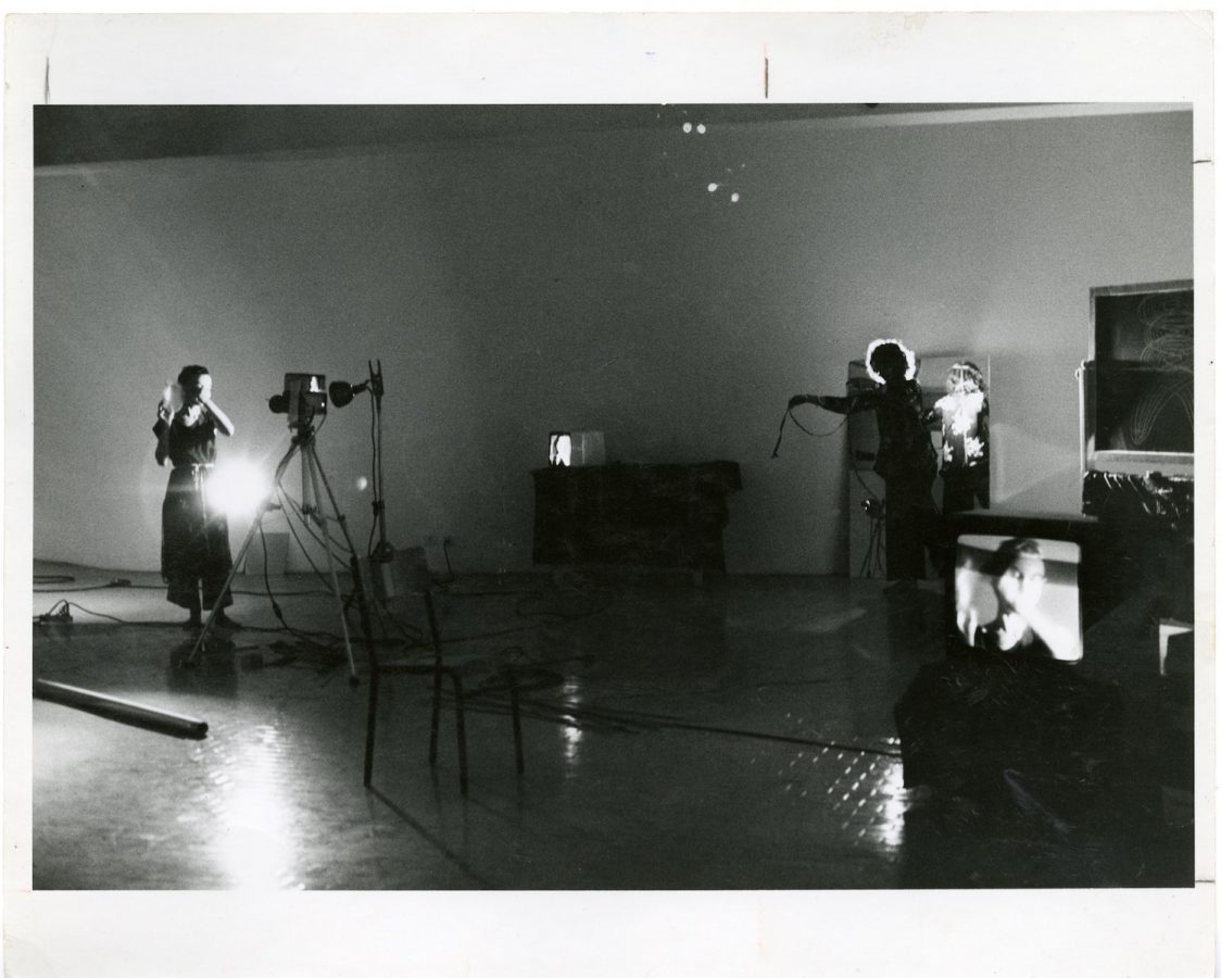 One performer stands in front of a camera while another stands in front of a mirror