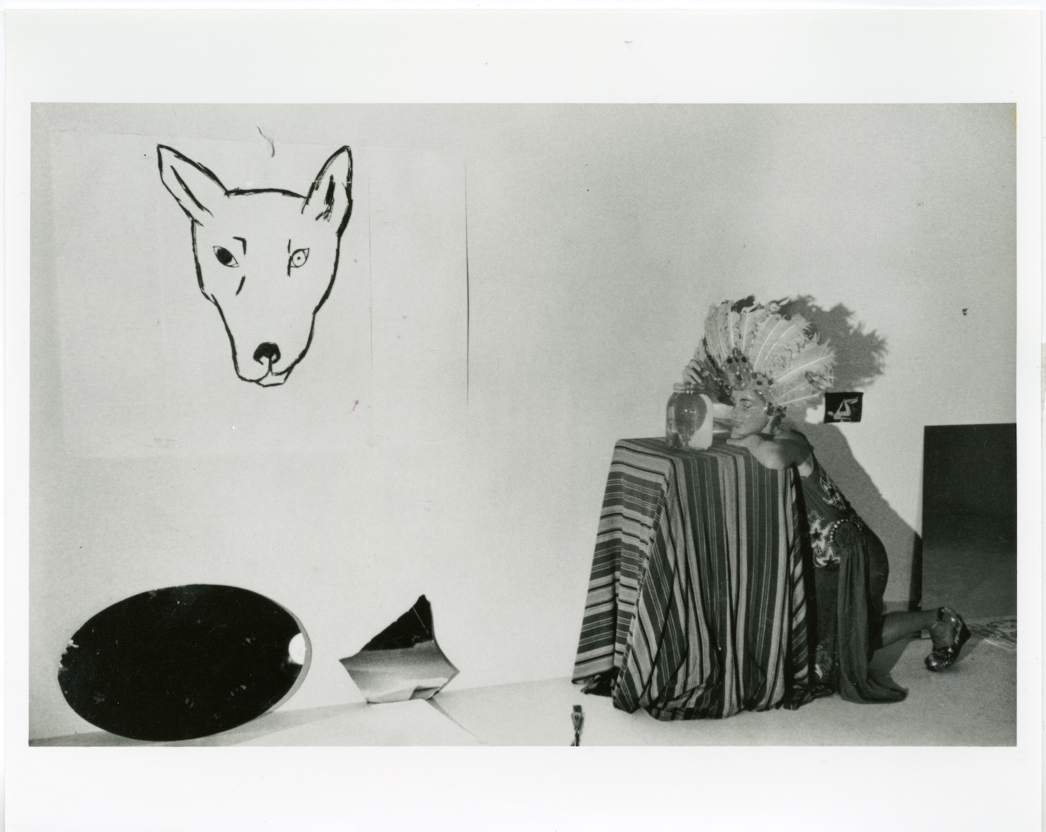 Jonas wearing a mask and feather headpiece while leaning against a table and looking into a jar of water. On the wall is a drawing of a dog's head.