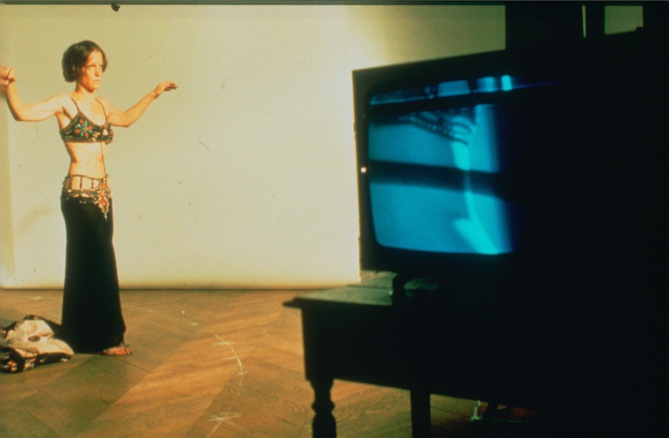 Jonas stands with her arms extended to the side while dressed in a bra and long skirt, a video monitor is to the right.