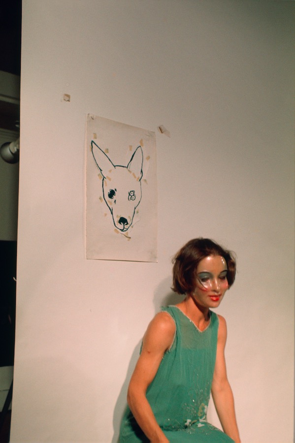 Jonas wearing a mask and a green dress, sitting under a drawing of a dog’s head