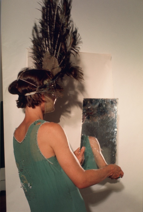 Jonas looking into a mirror while wearing a peacock-feathered headdress, mask, and green dress