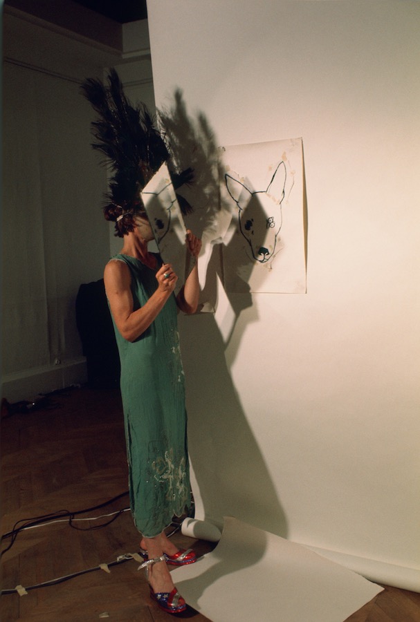 Jonas standing next to a drawing of a dog’s head and looking into a mirror while wearing a peacock feathered headpiece, mask, and green dress.