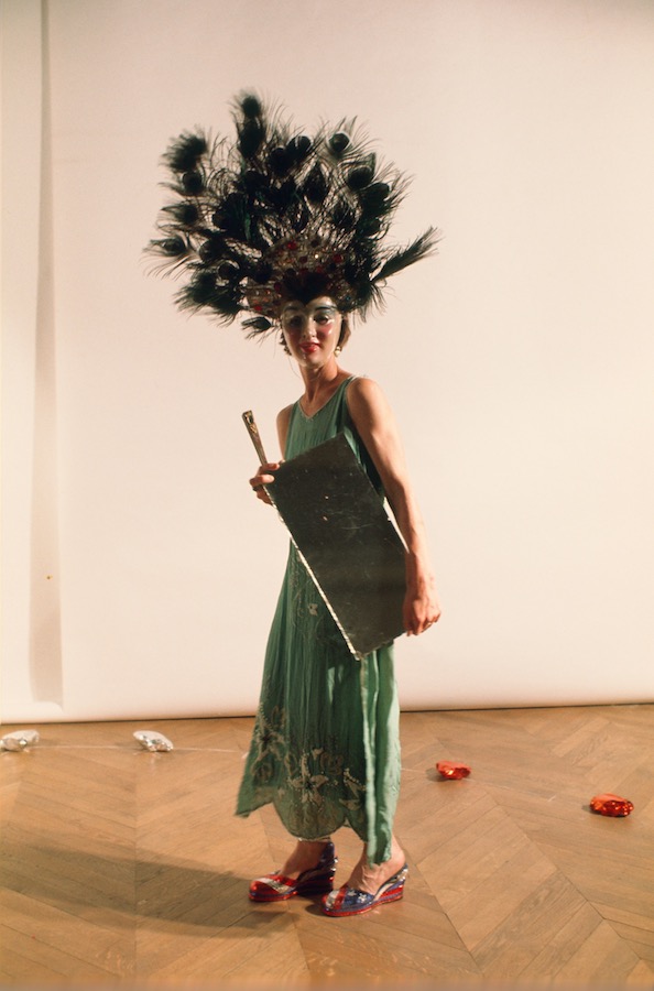 Jonas holding a mirror and fan, while wearing a peacock-feathered headpiece, mask, and green dress