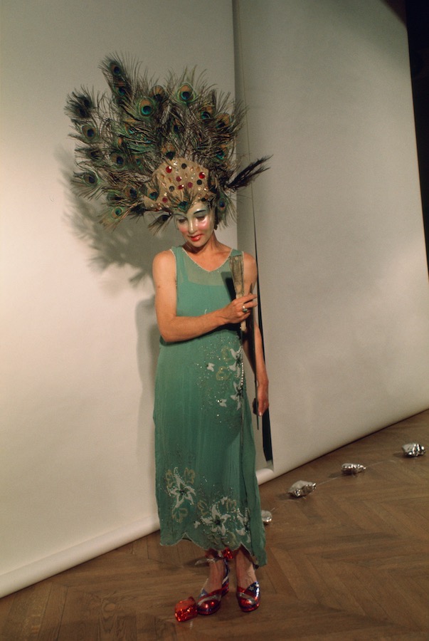 Jonas holding a mirror and fan, while wearing a peacock-feathered headpiece, mask, and green dress