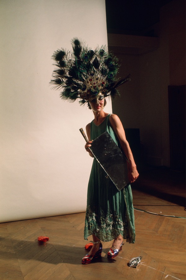 Jonas holding a mirror and fan, while wearing a peacock-feathered headdress, mask, and green dress