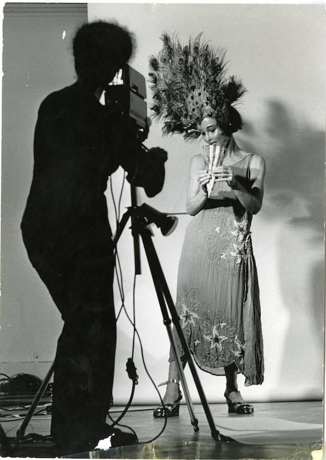 Jonas holds a fan while standing in front of a camerawoman and wearing a peacock-feathered headpiece, mask, and dress.