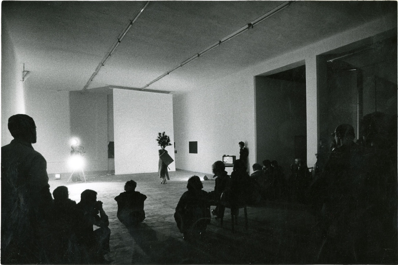 Audience watching Jonas holding a mirror while wearing a peacock-feathered headdress and dress