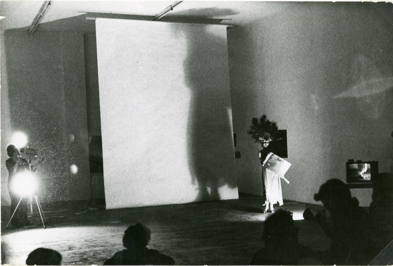 The audience watches Jonas perform in front of a white backdrop while wearing a peacock-feathered headpiece and dress and holding a mirror. There is a camerawoman to the left and a video monitor to the right.