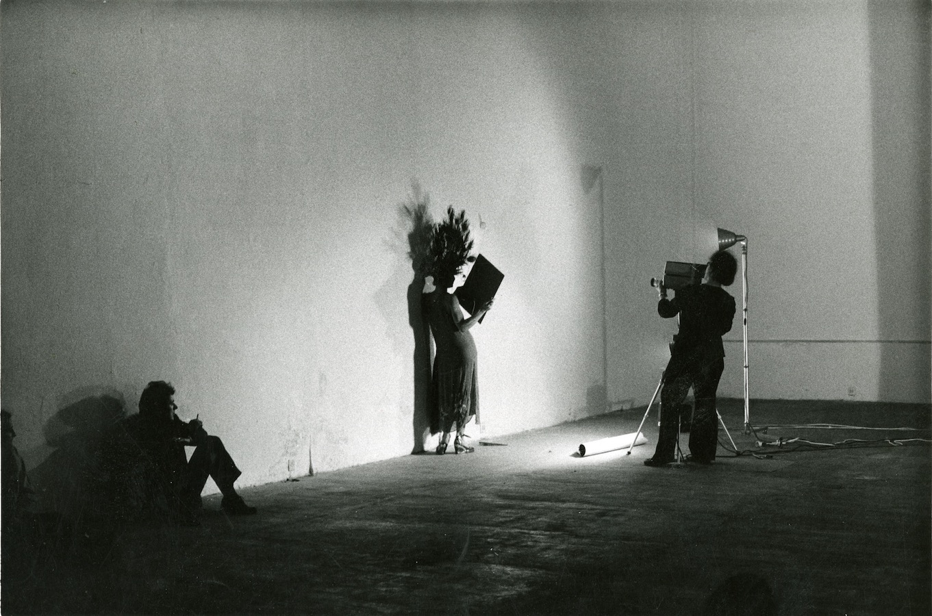 A camerawoman films Jonas holding a mirror while wearing a peacock-feathered headpiece and dress. To the left of Jonas, a member from the audience watches from the floor.