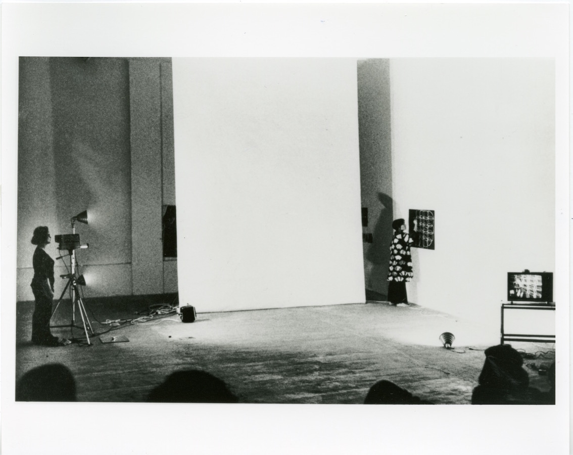 Camerawoman films Jonas drawing on a paper against the wall while a video monitor displays the recording to the audience.