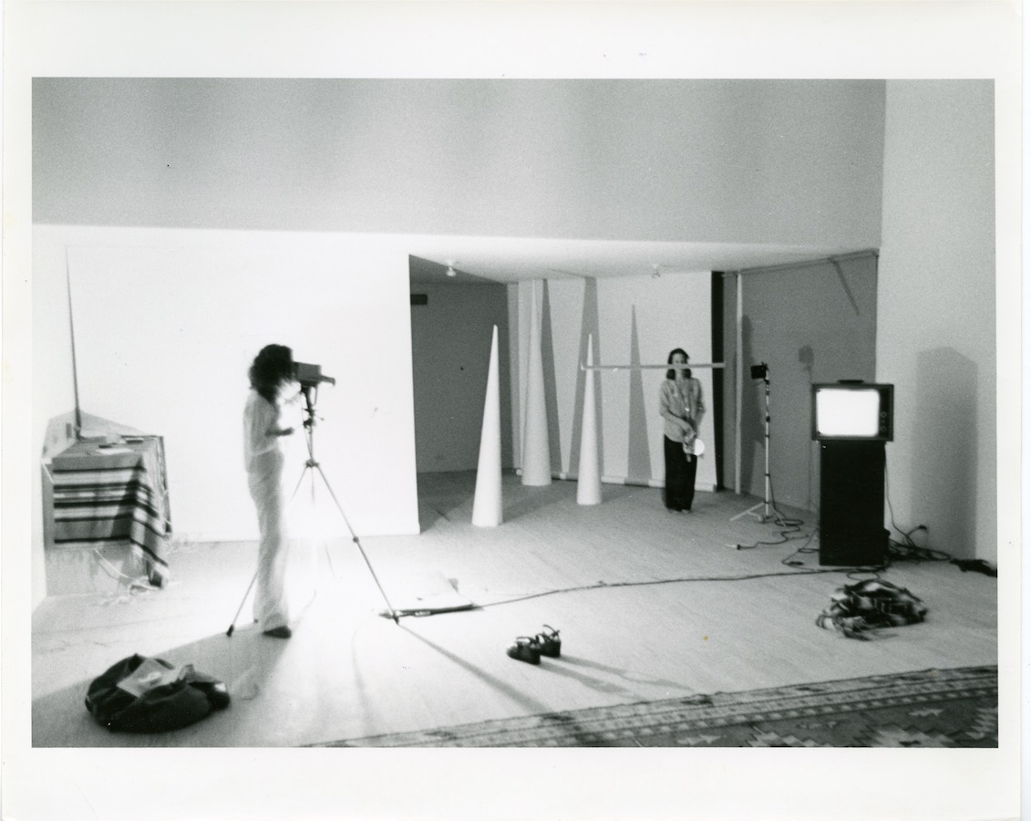 A camerawoman stands behind a tripod to film Jonas standing behind a pole that is attached to a white cone. Two more white cones and a video monitor on a stand are also in the room.