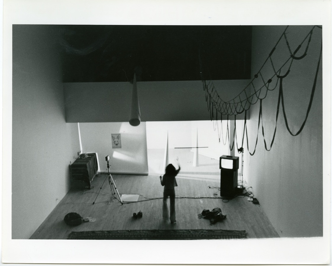 View from the mezzanine level: a fabric artwork hangs from the ceiling and an unseen performer holds a white cone hang from the mezzanine. On the ground level, Jonas stands facing towards two white cones and a drawing of a dog’s head; to her left is a camera on a tripod, a mirror on the floor, and to her right is a video monitor on a stand.