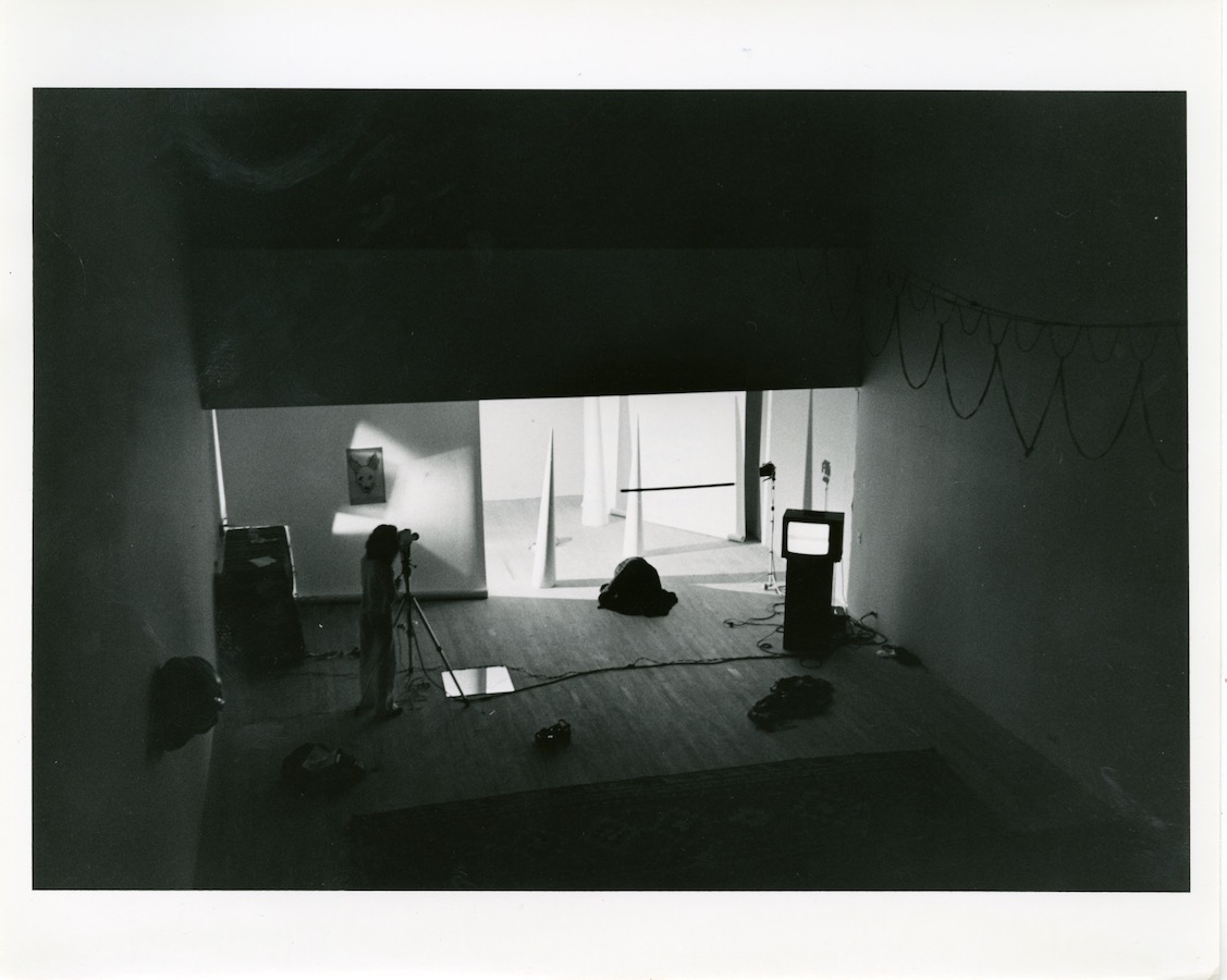 View from the mezzanine level, a camerawoman films Jonas peaking around the corner. The room is filled with two white cones, a mirror on the floor, a video monitor on a stand, and a drawing of a dog’s head.