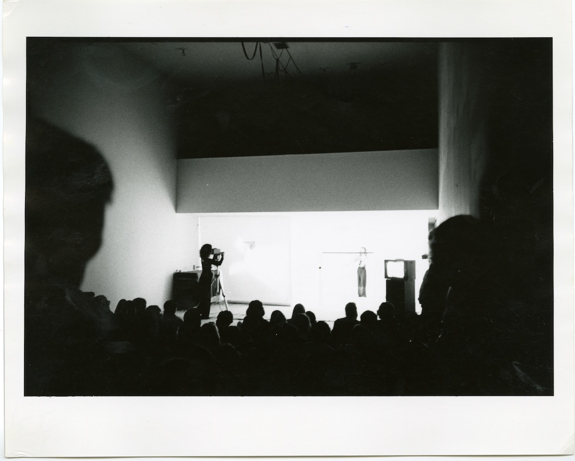 Crowd watching a camerawoman film Jonas as she performs behind a pole that is attached to a white cone. A video monitor on a stand is located towards the right.