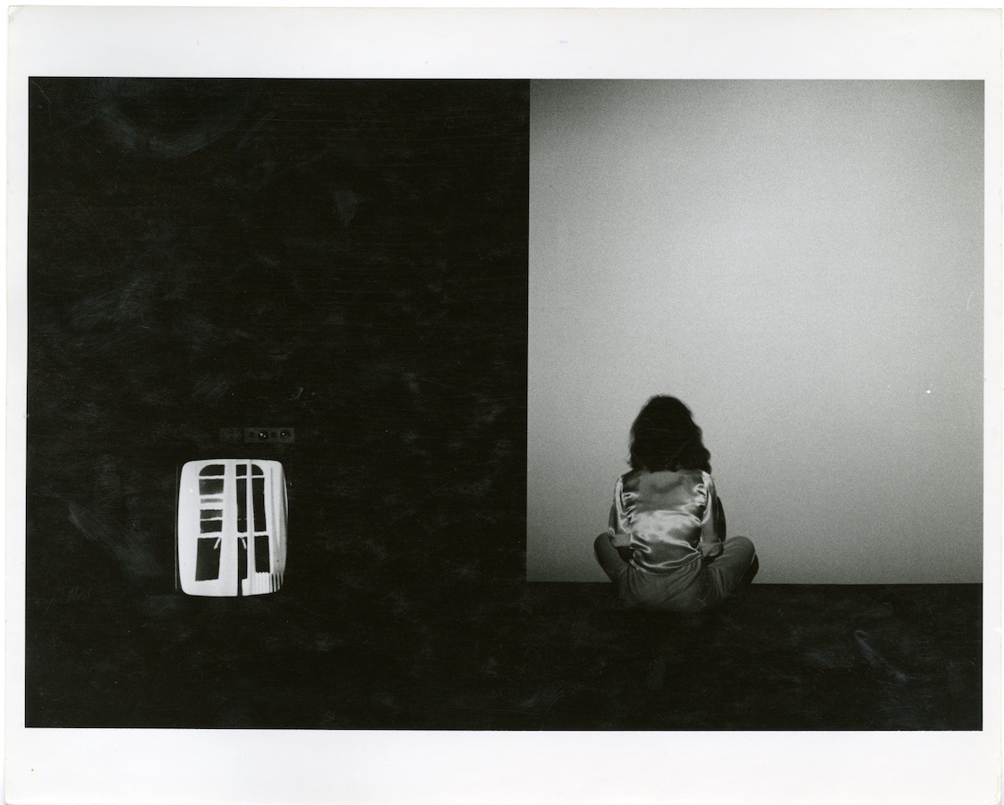 Jonas sits on the floor with her back to the camera; to the left is a TV monitor displaying a window.