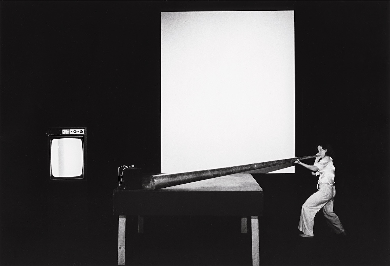 Jonas blowing into a large cone that is placed on a stage; to the left is a TV monitor.