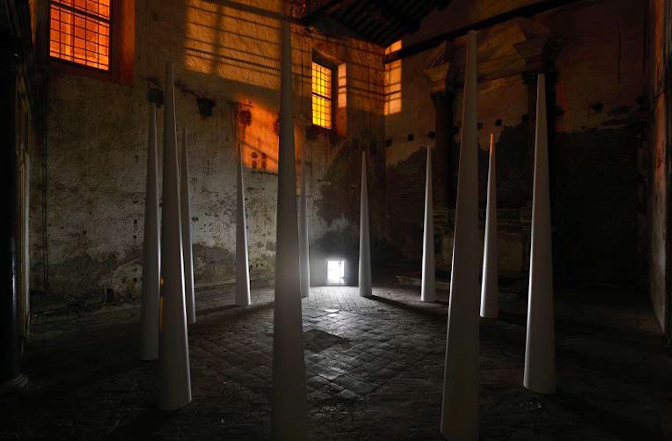 A circle of large cones, outside the circle is a bright TV monitor on the floor, above the cones are two windows casting light and shadows against the walls.