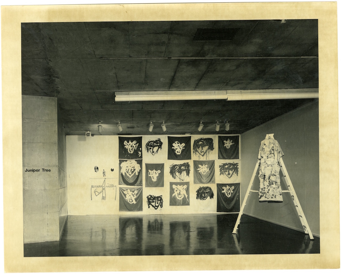 Various paintings hang against the back wall, to the right is a ladder with a robe hanging from it and to the left is a sign that reads: “Juniper Tree”