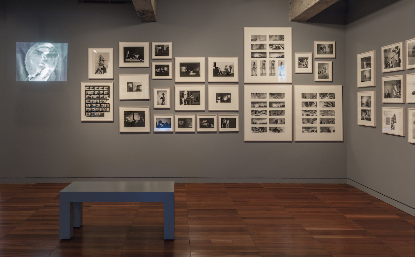 Various framed photographs and photographic contact sheets and a projection from Jonas’s performances are displayed on a wall. There is a bench in the middle of the room.