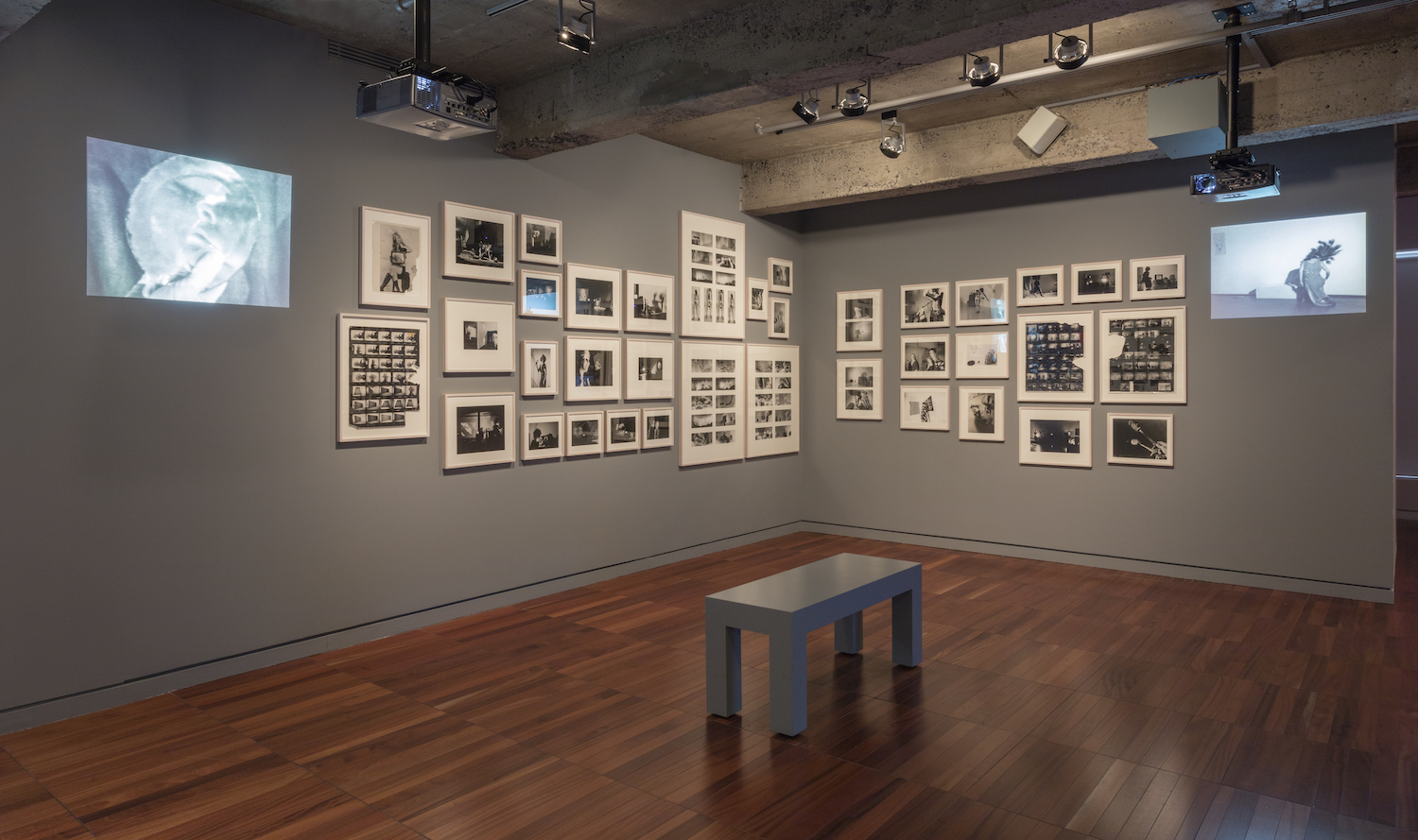 Various framed photographs and photographic contact sheets and two projections from Jonas’s performances are displayed on a wall. There is a bench in the middle of the room.