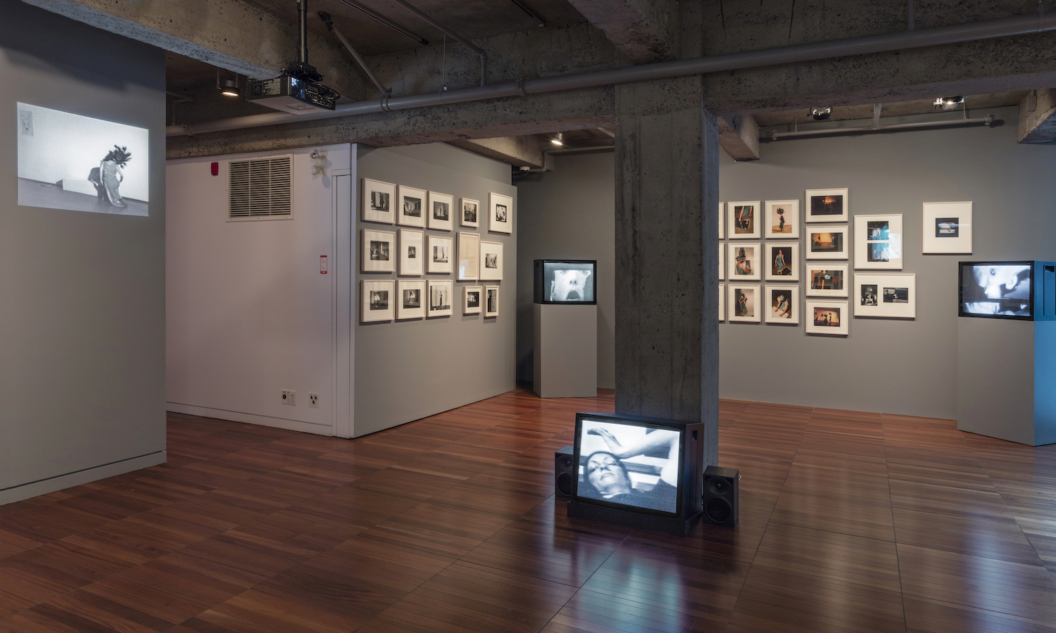 Various photographs and a projection are displayed on a wall. There are also three TV monitors, two on stands, and one on the floor.