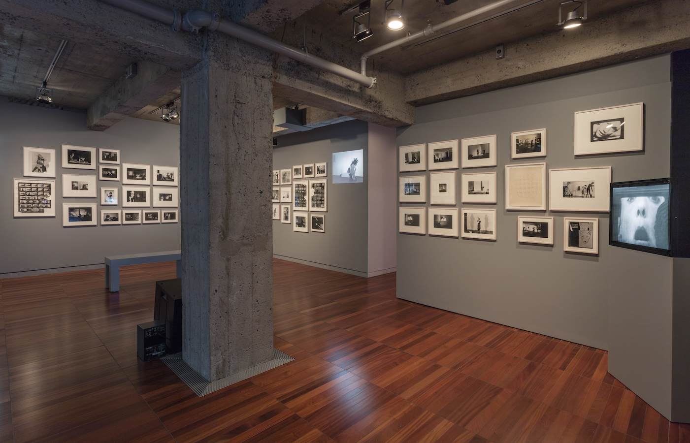 Various photographs and a projection are displayed on walls. There are also two TV monitors, one on a stand, and one on the floor, and a bench.