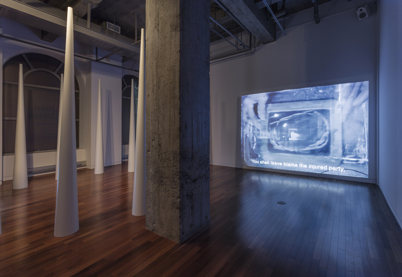 A large pillar separates the gallery; to the left is a circle of large cones and to the right is a video projection. The video projection reads: “You shall leave blame the injured party,”