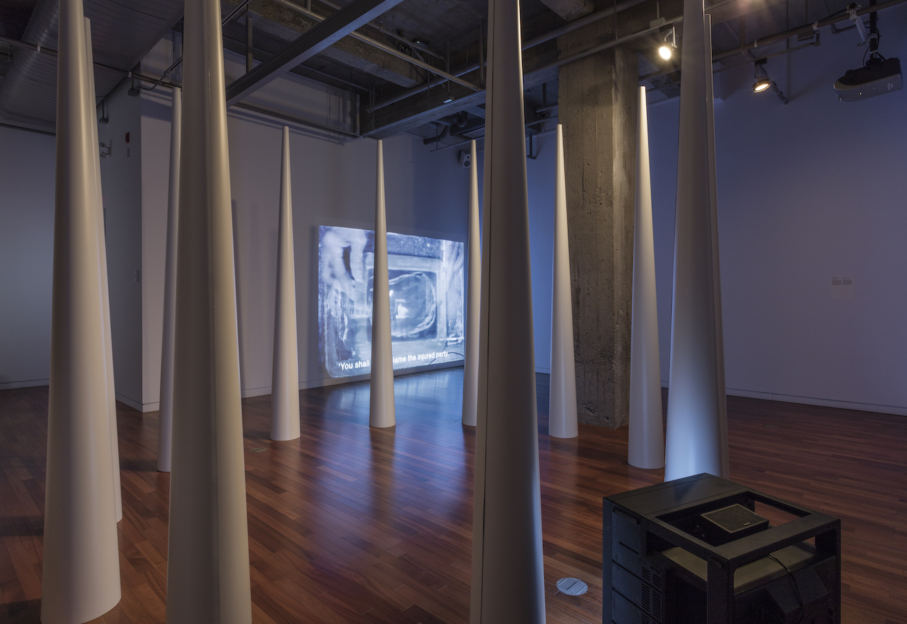 A circle of large cones; to the right is a TV monitor on the floor and in the background is a video projection.