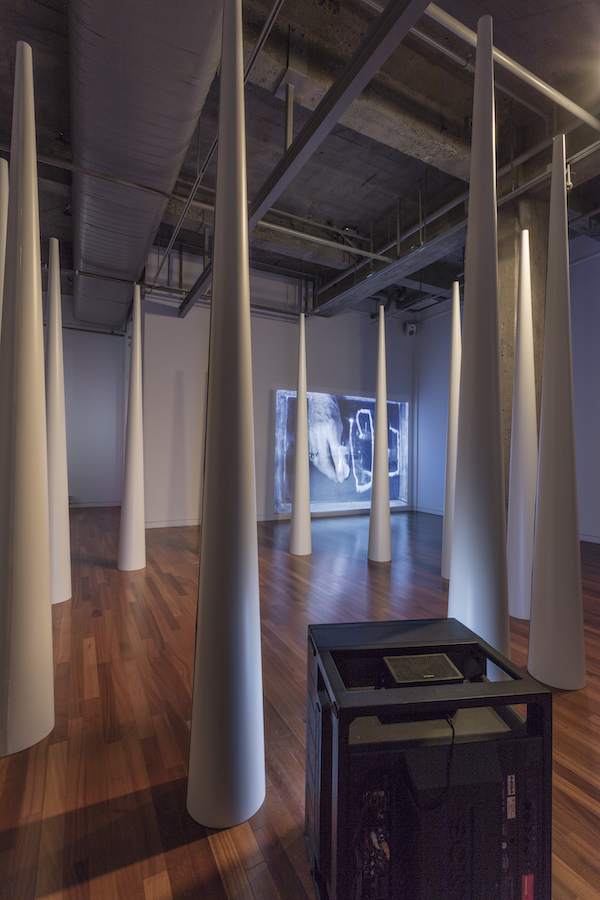 A circle of large cones; in the foreground is a TV monitor on the floor and in the background is a video projection.