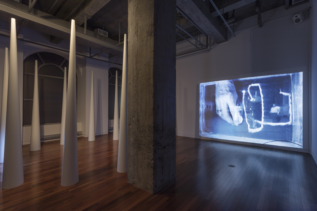 A large pillar separates the gallery; to the left is a circle of large cones and to the right is a video projection.