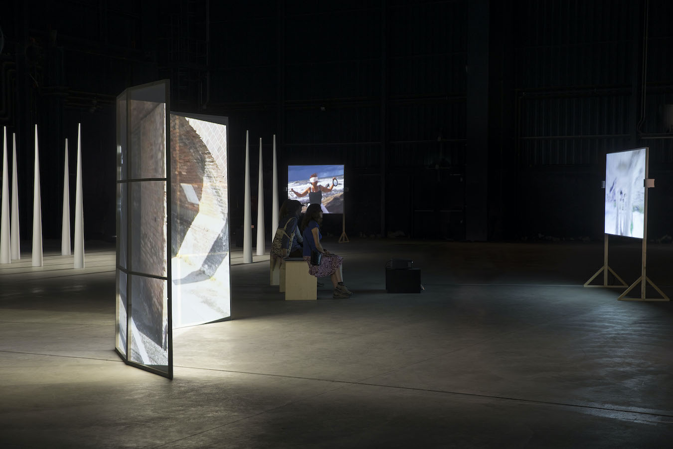 A figure sits on a bench to watch a video projection, in the background are large cones in a circle.