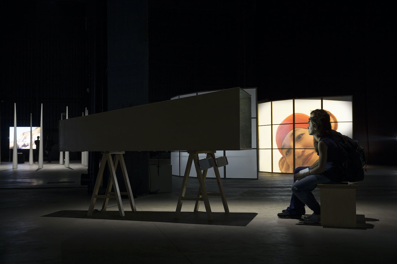 Two figures sit on a bench in front of a mini theater; in the background are video projections on curved screens and a circle of large cones.