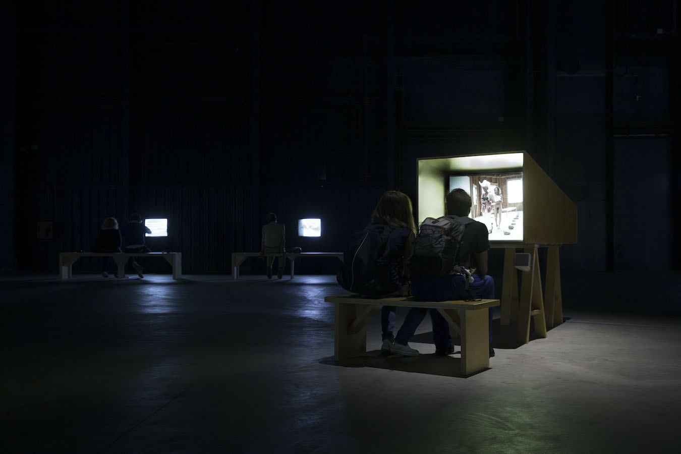 Two figures sit on a bench to watch a TV monitor, in the background are two more benches and TV monitors.