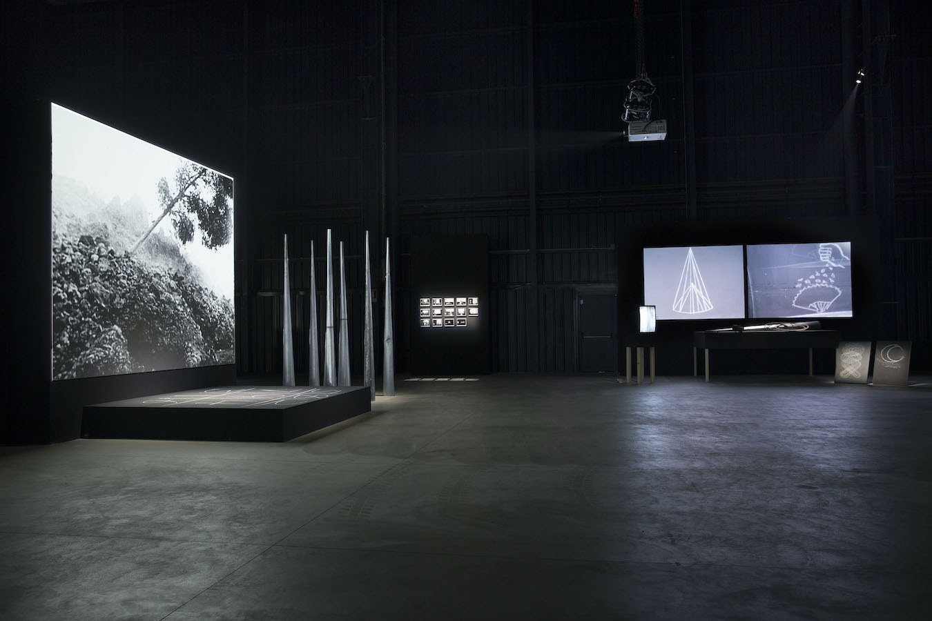 To the left are a circle of large cones next to a small stage with a number grid written in chalk; behind the stage is a projection of palm trees. To the right are two projections of chalk drawings, a TV monitor on a stand, a table with a hoop and large cone placed on top, and two chalk drawings on the floor. Framed photographs from Jonas’s performance hang on the back wall.