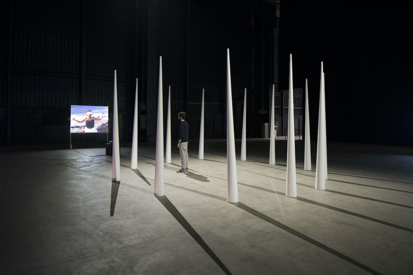 A figure stands in a circle of large cones facing a video projection.