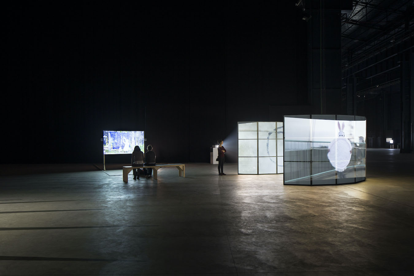 Two figures sit on a bench to watch a video projection, to the right is a standing figure viewing two video projections on curved screens.