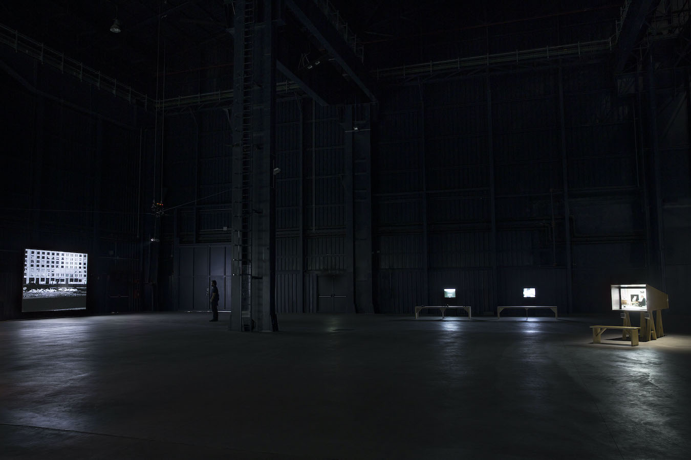 A dimly lit room filled with a video projection to the left and three TV monitors with benches to the left.