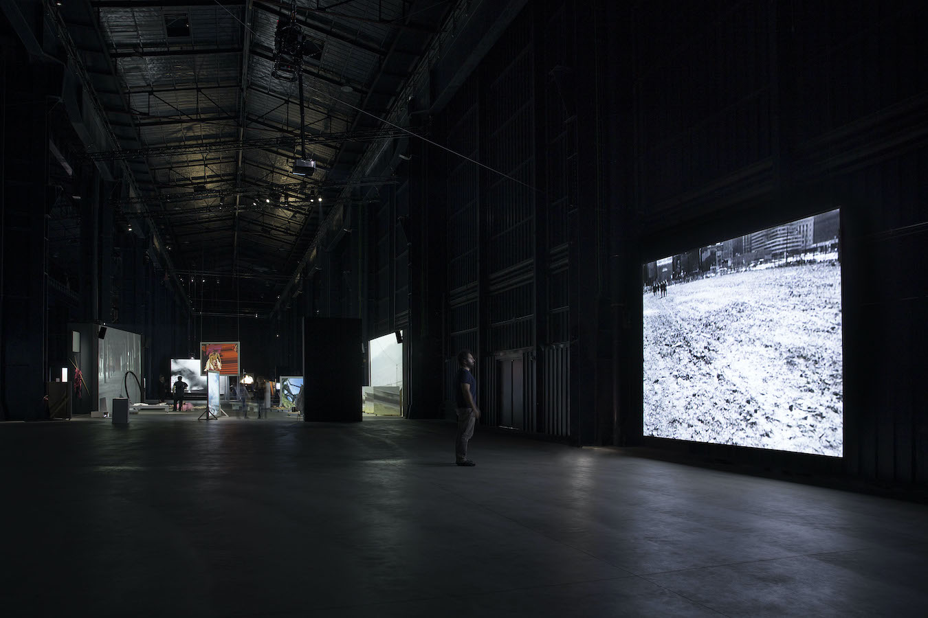 A figure standing in front of a video projection, in the background are various props and projections.