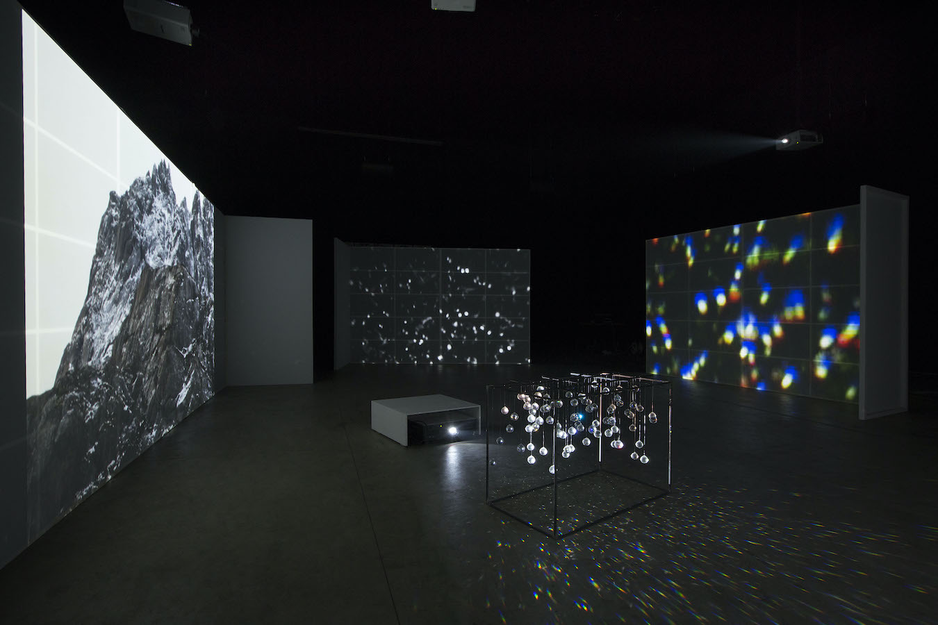 Three video projections surround a projector placed on the floor, illuminating various crystal orbs hanging from a frame. The crystals are casting reflections across the floor.