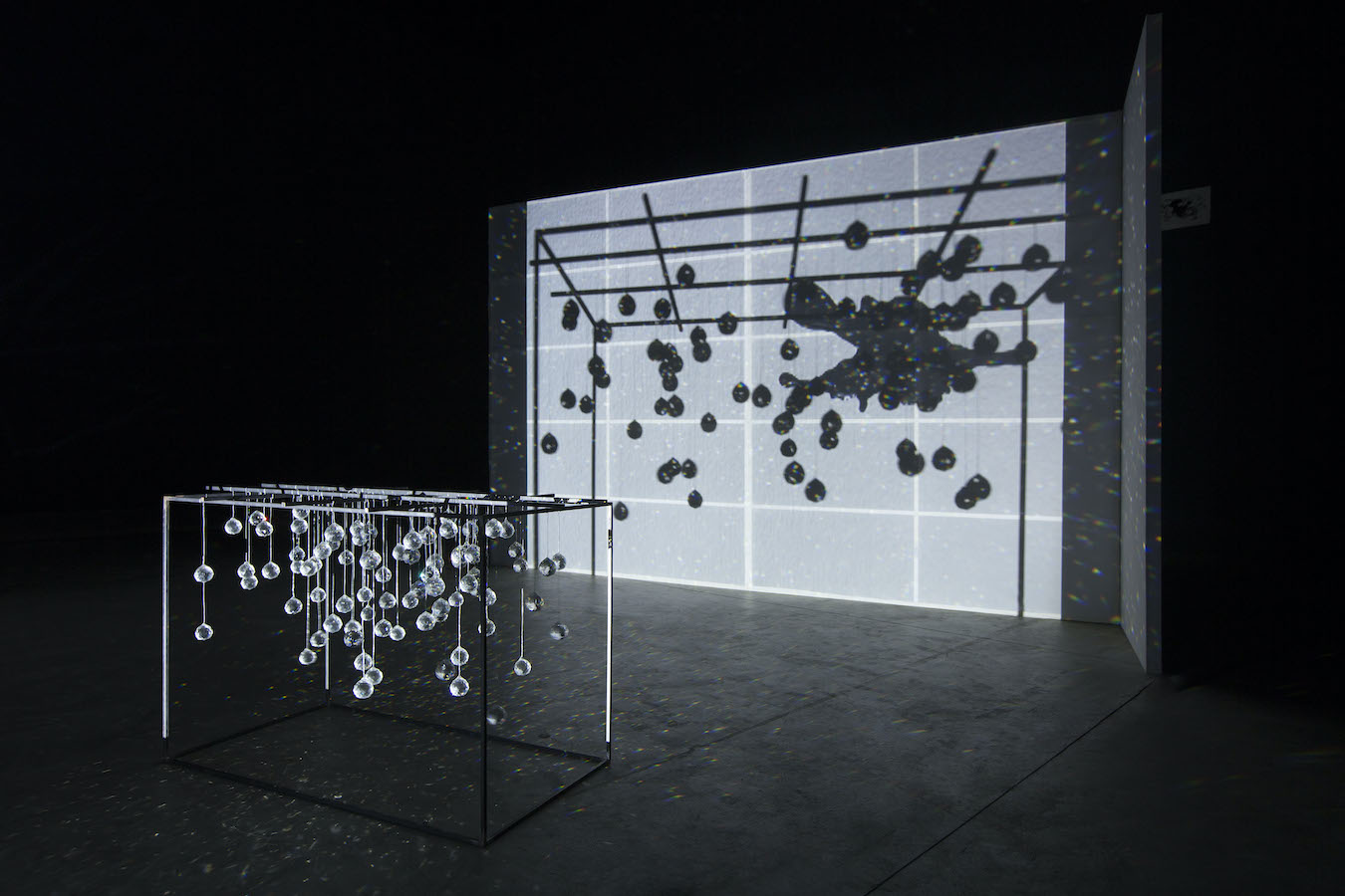 A sculpture made of hanging crystals is placed on the floor and is casting reflections, behind it is a video projection.