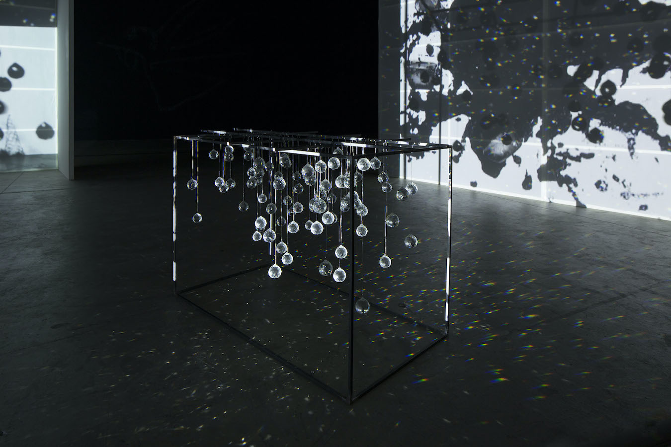 A sculpture made of hanging crystals is placed on the floor and is casting reflections, surrounding it are video projections