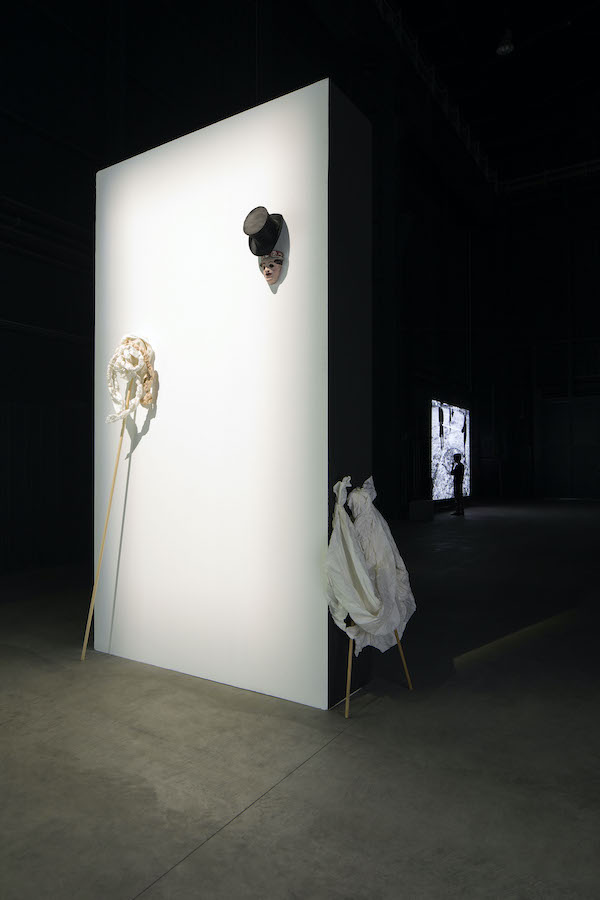 Various sculptures lean against a wall, in the background is a figure standing in front of a video projection