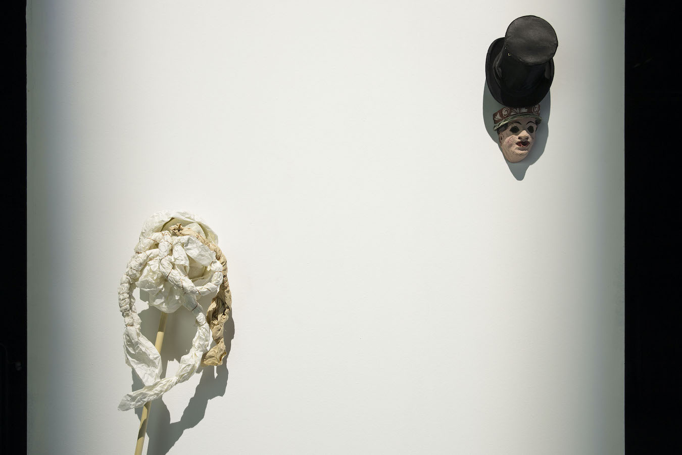 To the left is a sculpture of a mop leaning against a wall, to the left is a sculpture of a face and a top hat hanging on the wall