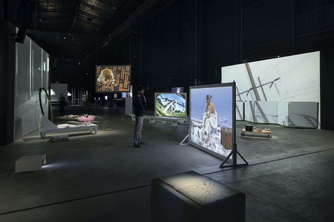 A figure stands in a room surrounded by various video projections and props