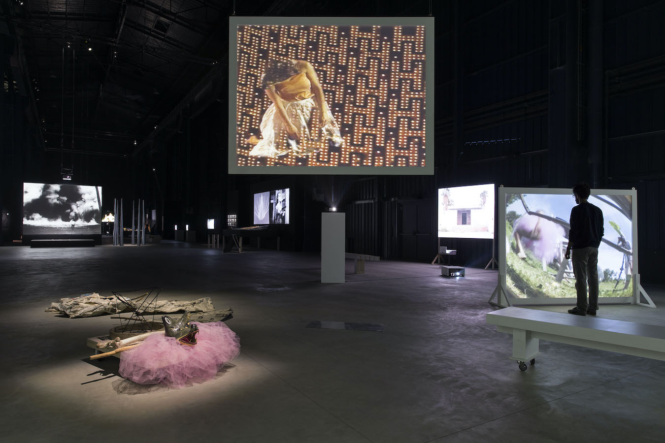 A room filled with various video projections and props, to the right is a figure standing in front of a video projection