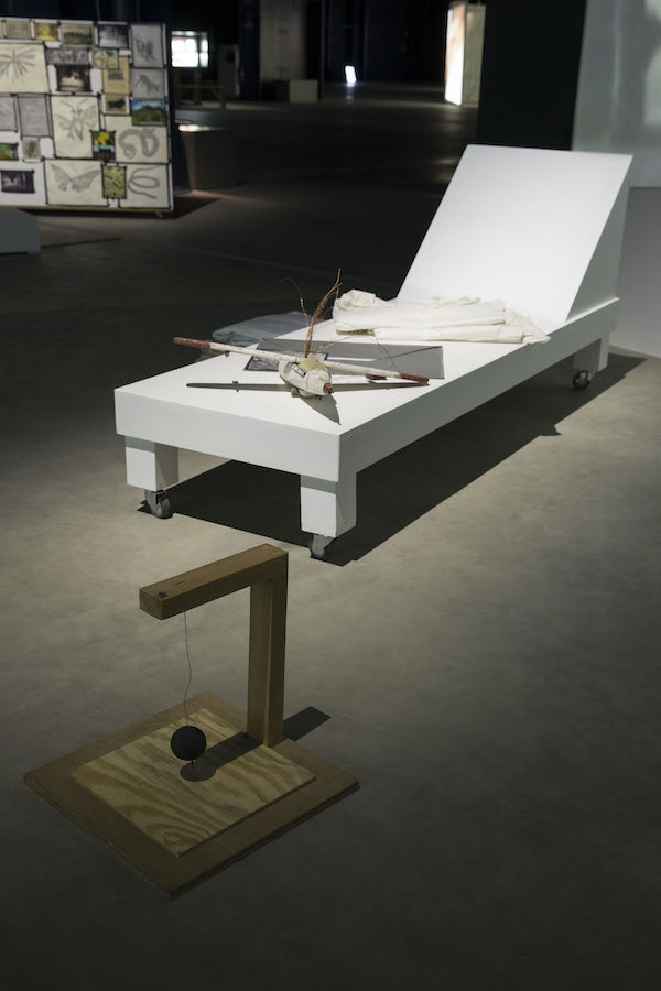 A pendulum is placed on the floor, behind it is a lounge chair with various props on top of it.