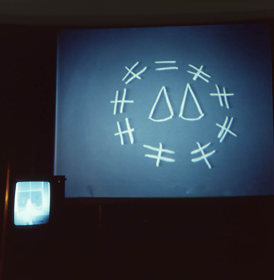 Projection of a chalk drawing of two cones in the center of a circle made up of various lines on a blackboard. In the left corner is a TV monitor projecting an image of a figure in front of a window.