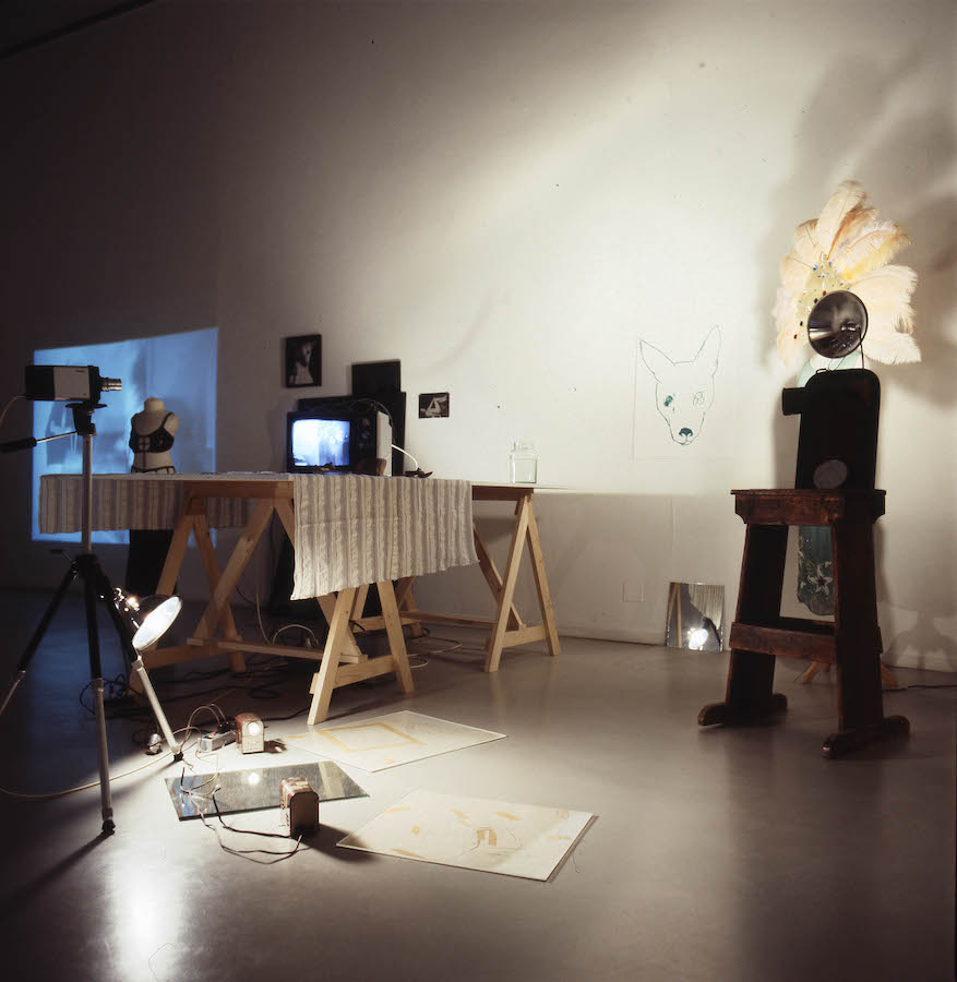 A camera on a tripod faces various props used for Jonas’s performance, a light is attached to the tripod and is pointed to the floor. There is a TV monitor and a glass jar placed on a table draped with a cloth; on the wall there is a projection to the far left, various images in the middle, and a drawing of a dog’s head with four eyes to the right. To the right of the drawing is Jonas’s dress and peacock-feathered headpiece on a mannequin, which is illuminated by a light placed on a table in front of the clothing. On the floor are various mirrors and drawings.