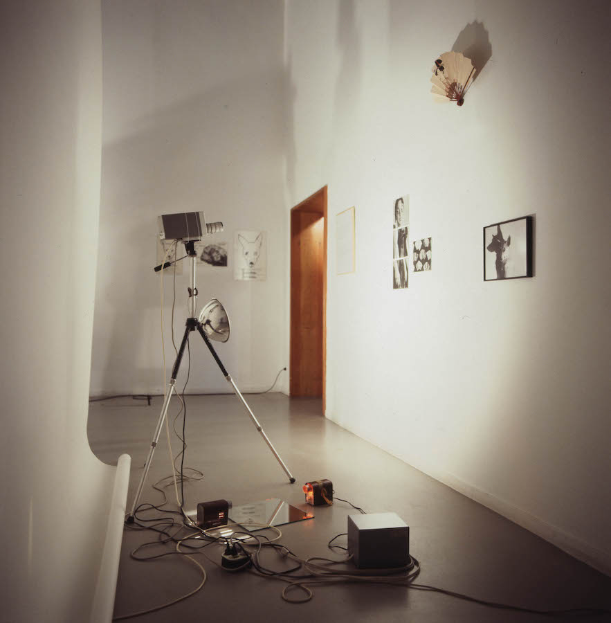 A light and camera attached to a trip pod are pointed towards a wall with various photographs hanging on it. Above the photos hangs a paper fan and on the far wall in the background are various drawings. Behind the tripod is a backdrop and there is a mirror on the ground. The light attached to the tripod is creating dramatic shadows.